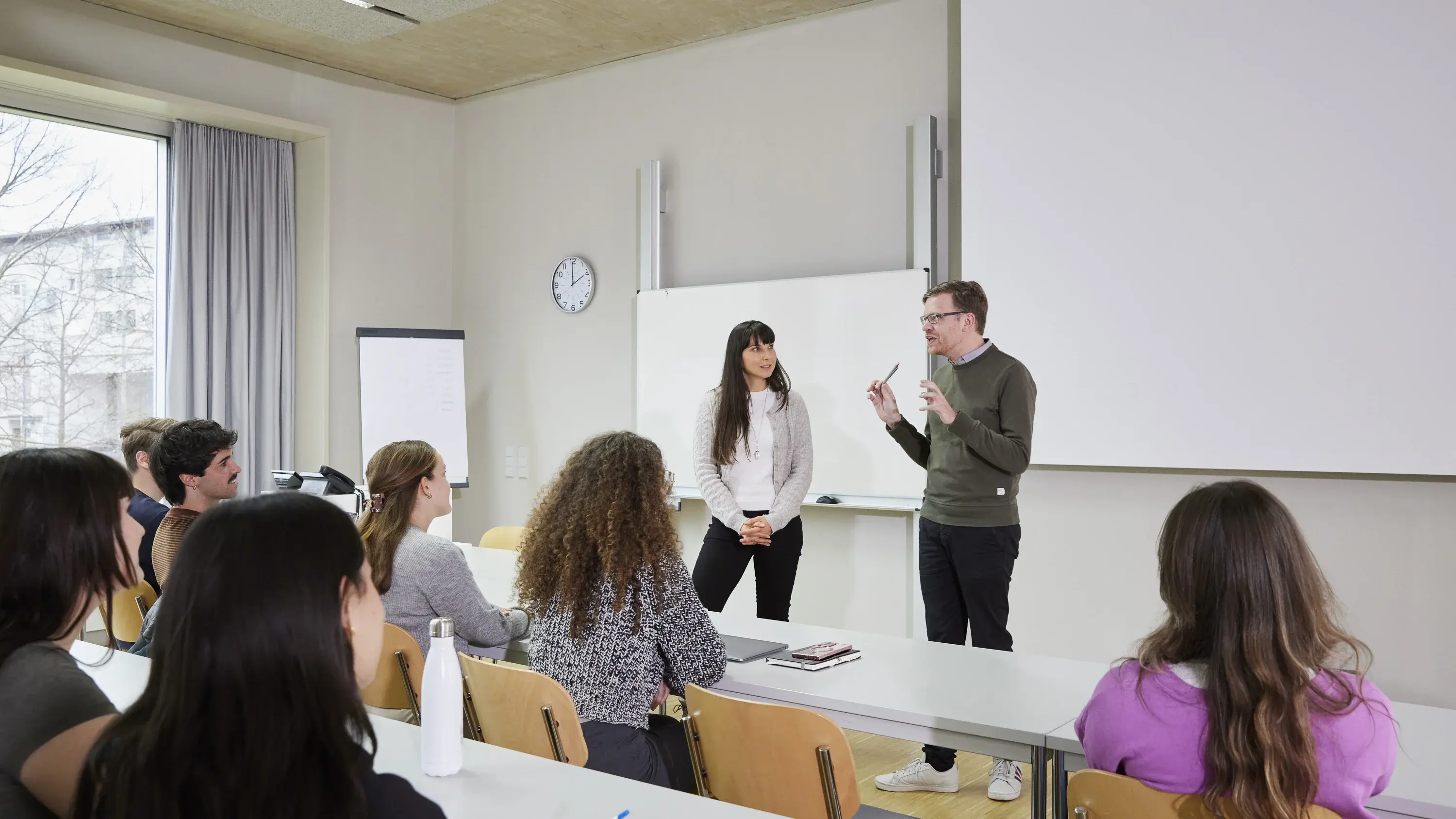 Eine Studierende und ein Professor sprechen in einer Vorlesung bei dessen Pult.