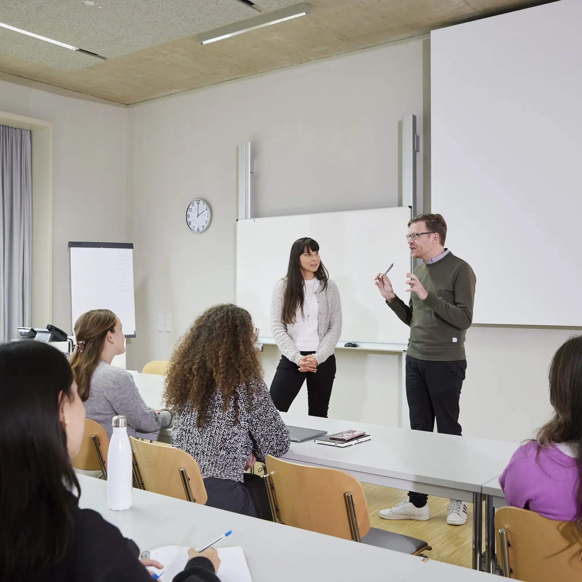 Eine Studierende und ein Professor sprechen in einer Vorlesung bei dessen Pult.