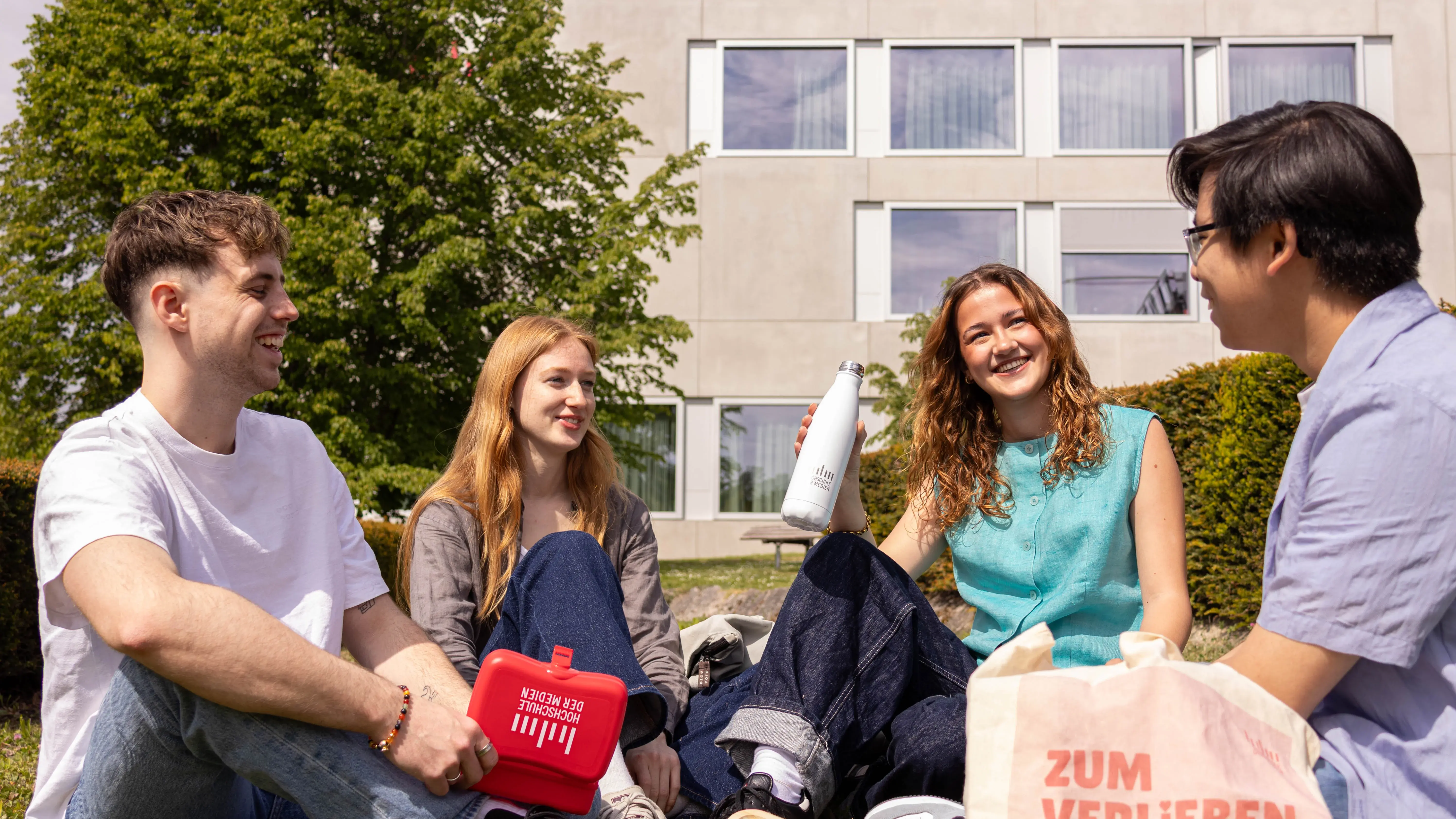 Studierende verbringen ihre Pause auf der Wiese des sonnigen Campus der Hochschule der Medien.