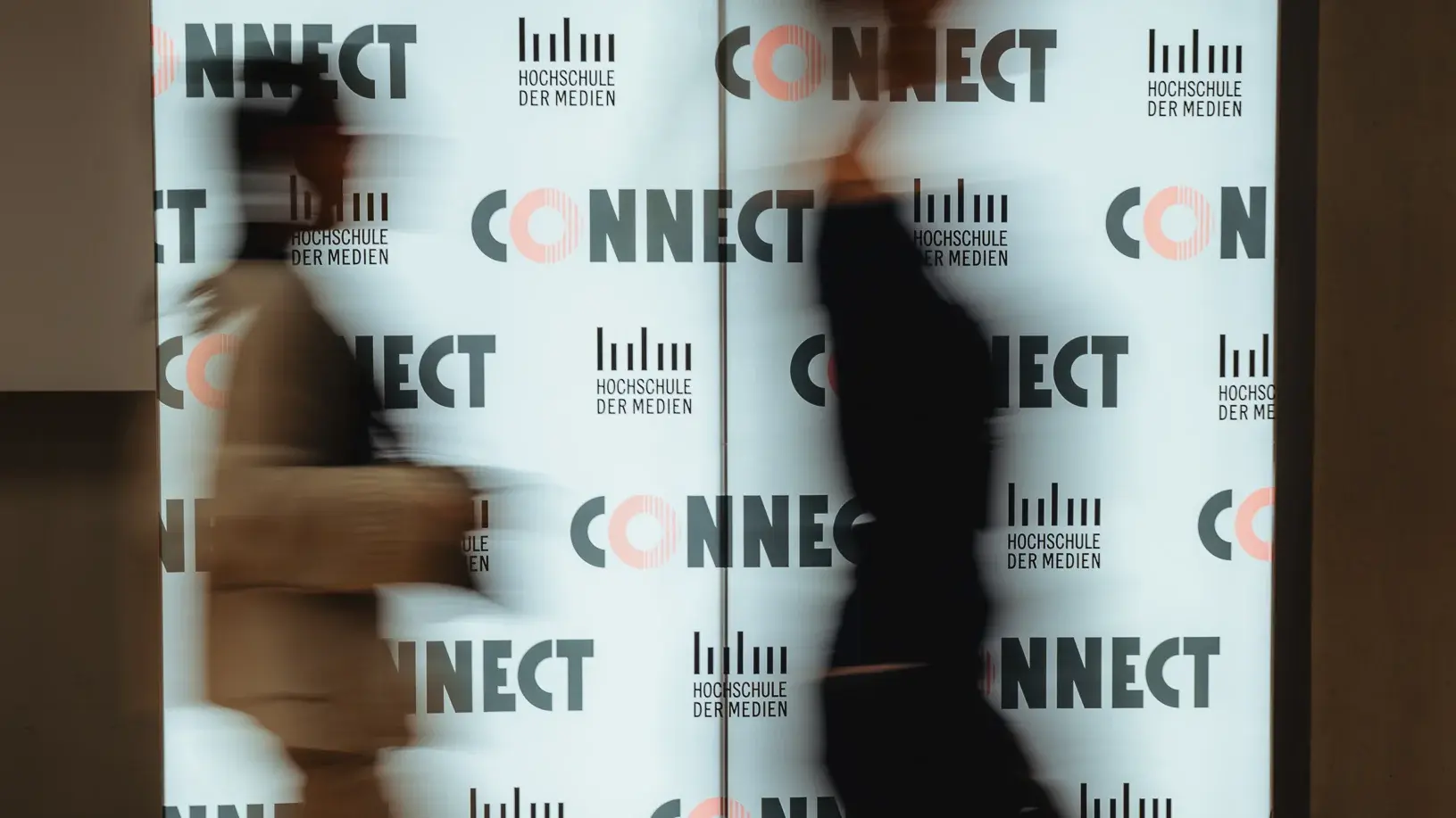 Zwei Alumni laufen beim Connectival 2025 an einer leuchtenden Fotowand vorbei.
