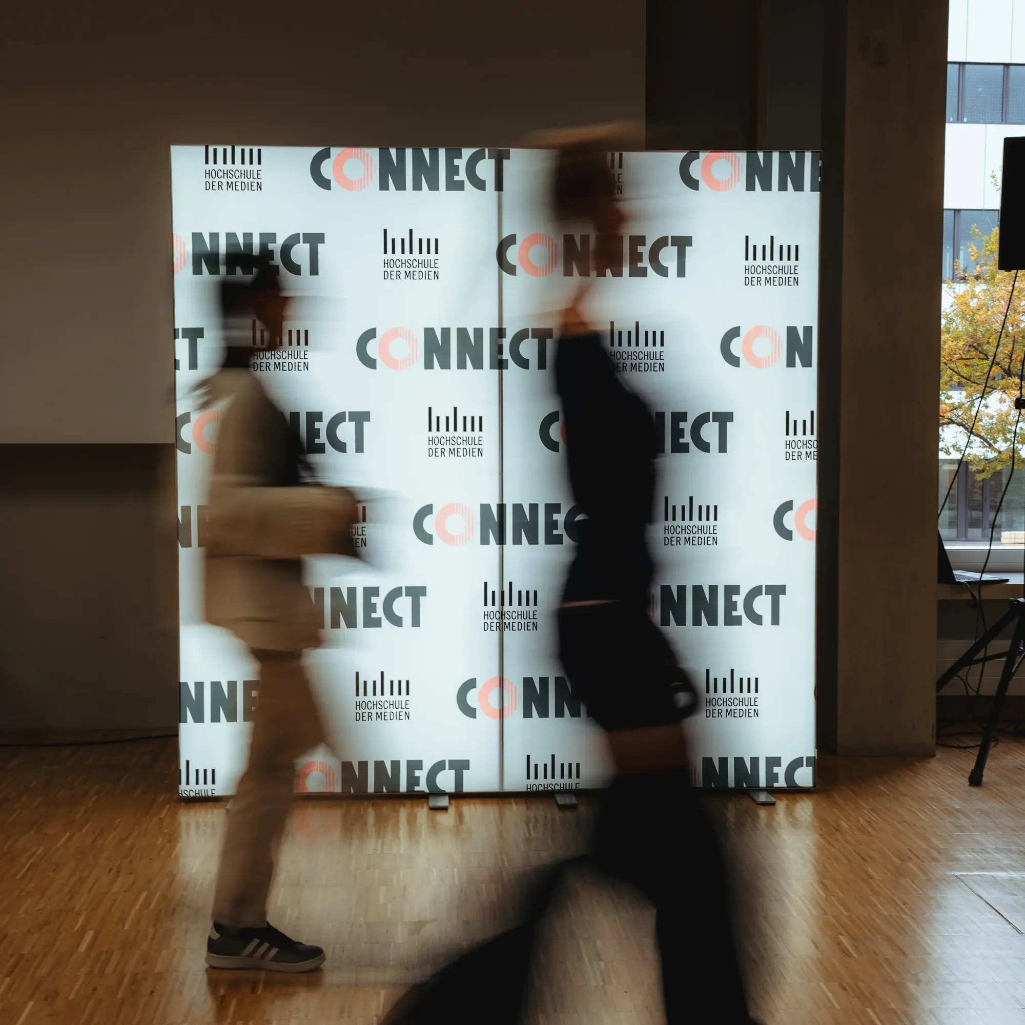 Zwei Alumni laufen beim Connectival 2025 an einer leuchtenden Fotowand vorbei.