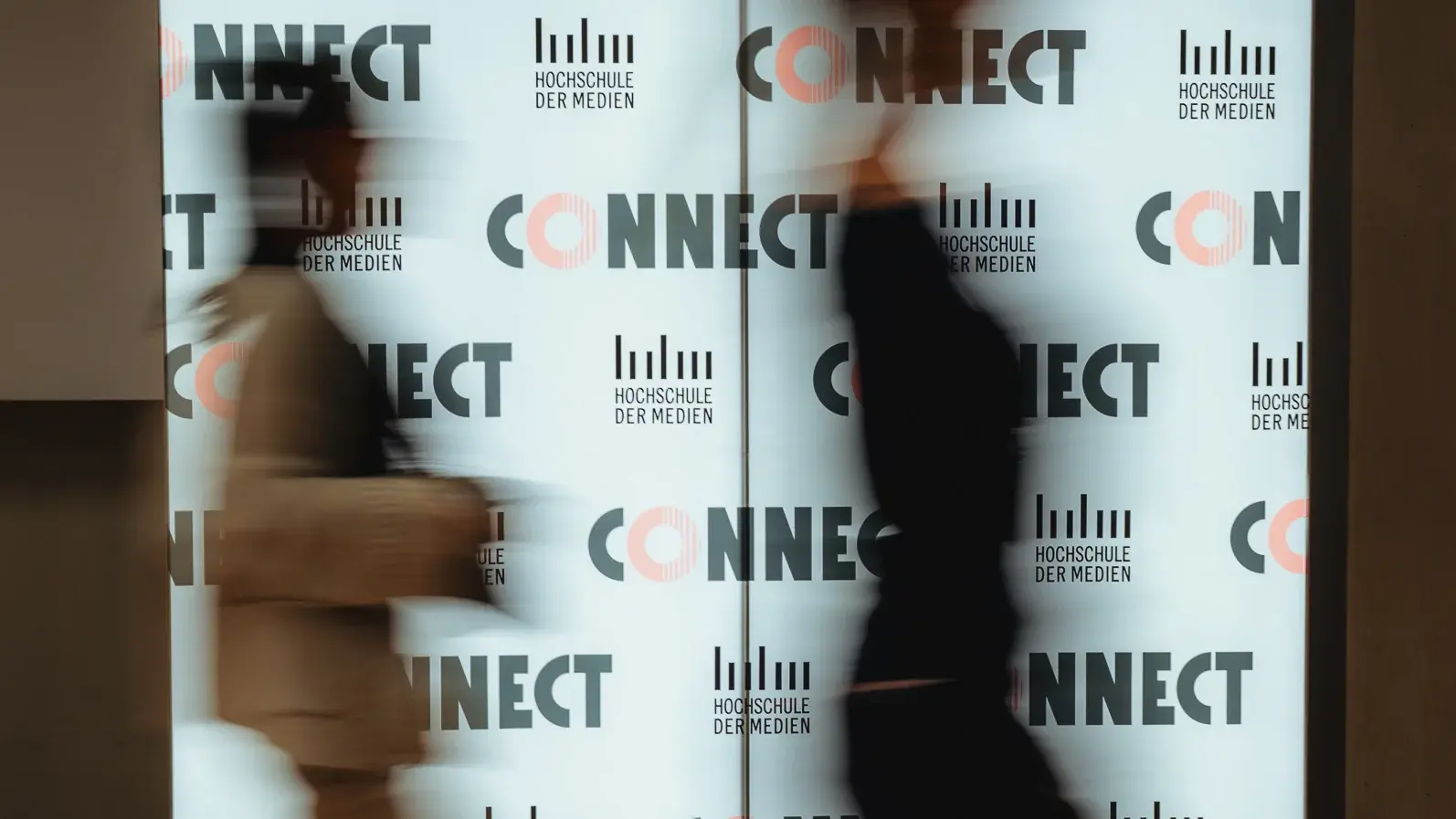 Zwei Alumni laufen beim Connectival 2025 an einer leuchtenden Fotowand vorbei.