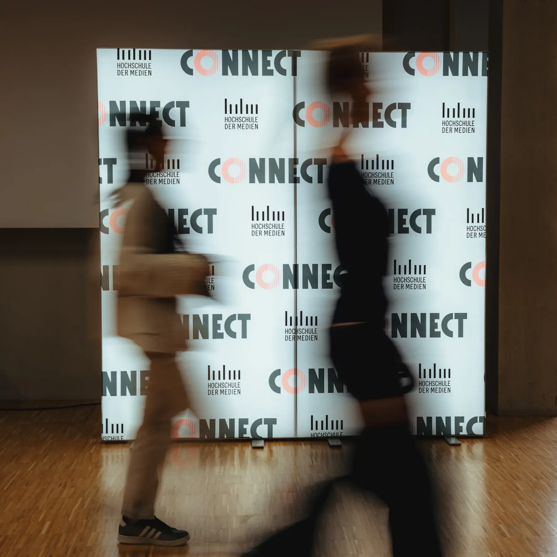 Zwei Alumni laufen beim Connectival 2025 an einer leuchtenden Fotowand vorbei.
